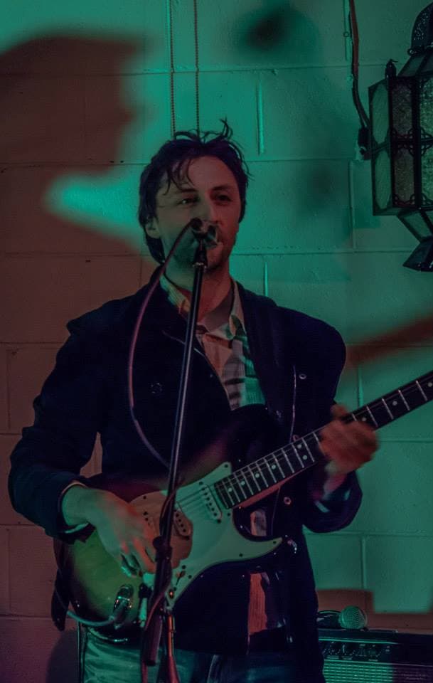 Ben sings into the mic while playing a Stratocaster guitar against a teal-lit brick wall at a gig.