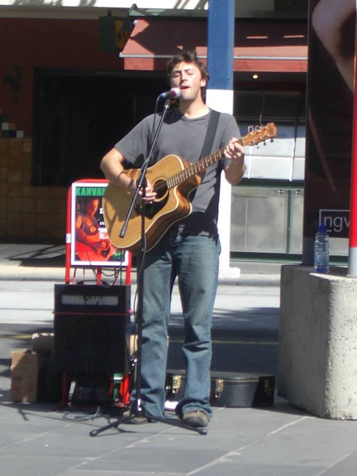 Ben busks on a sunny city street, singing into a mic while playing acoustic guitar near a Kanvas Grey CD stand.