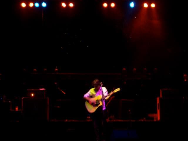 A lone musician plays acoustic guitar centre stage under colourful spotlights in a large concert hall.