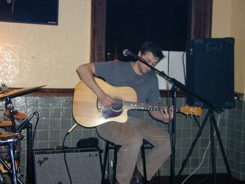 Ben sits on a stool playing acoustic guitar beside a speaker and drum kit during a quiet pub set.