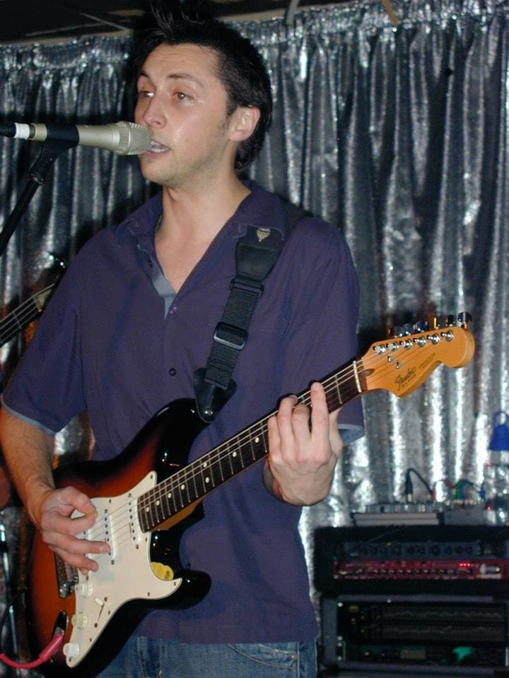 Ben sings into a microphone while playing a sunburst Fender Stratocaster in front of a silver curtain backdrop.