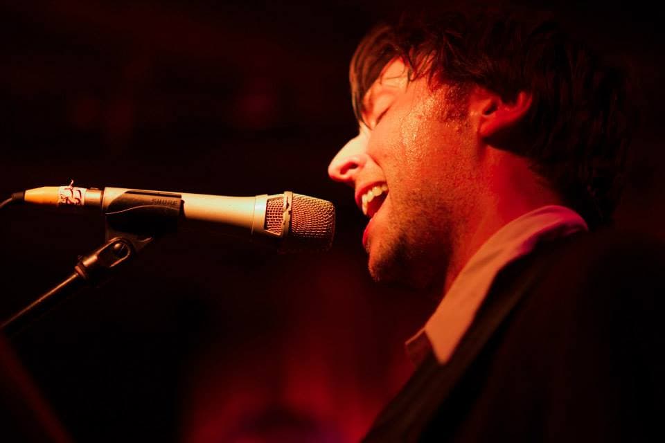 Profile close-up of Ben singing passionately into a Shure mic under deep red stage lighting.
