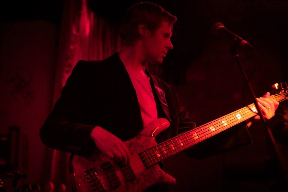 Liam plays his five-string bass under deep red stage lighting, leaning toward a vocal mic at a live show.