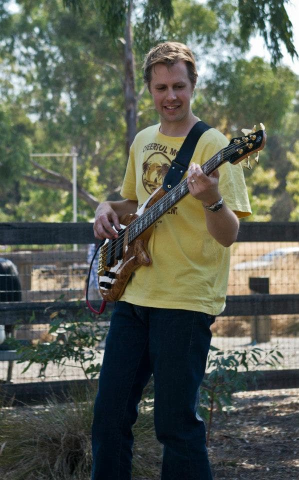 Liam smiles while playing a burl-top bass outdoors in a yellow t-shirt, with trees and a fence behind him.