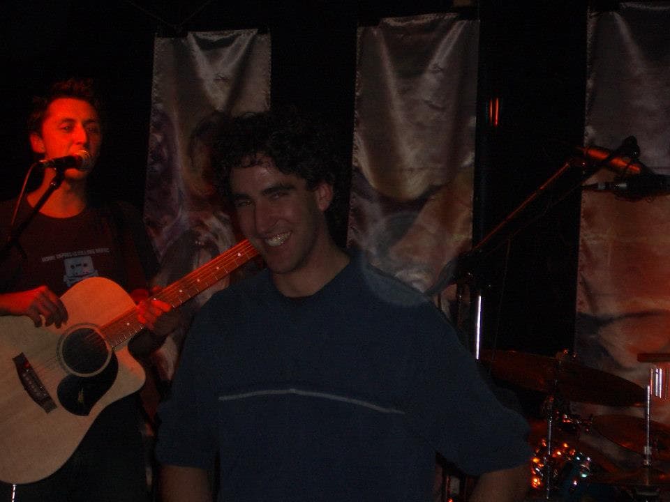 A grinning band member in a blue t-shirt stands at the front of a dim stage while Ben sings and plays acoustic guitar behind him.