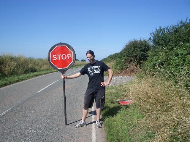 A smiling band member stands at the roadside on a sunny day holding a large red STOP sign on a pole.
