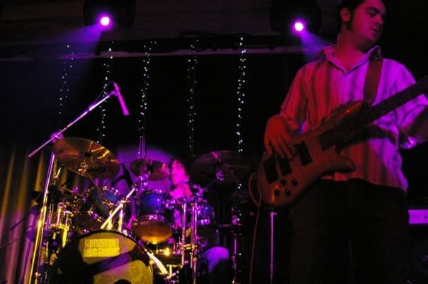 Justin plays bass on the right of a purple-lit stage while Mark drums in the background behind a curtain of fairy lights.
