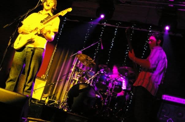 Kanvas Grey performing live on a fairy-lit stage with Ben singing and playing guitar, the drummer mid-song, and a bassist throwing his arms up.