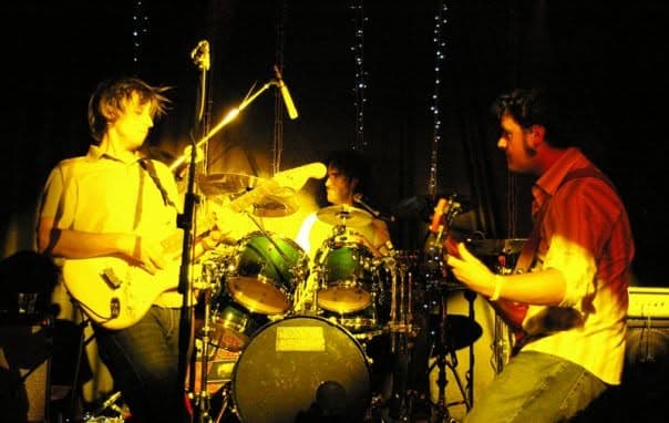 Kanvas Grey on a dim stage bathed in warm light, guitarist on the left, drummer at centre and a second guitarist leaning in on the right.