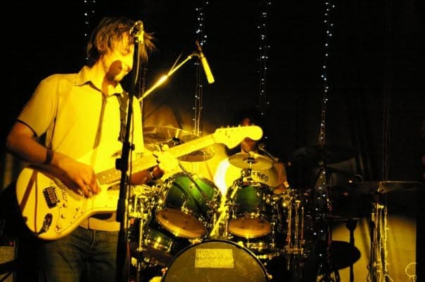 A guitarist sings into a mic while playing a Telecaster on stage, hair lit golden, with the drum kit glowing behind him.