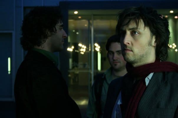 Three band members talk closely on a city street at night, lit by shop windows, one in a scarf in sharp profile.