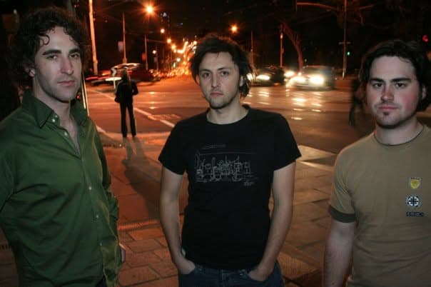 Three band members stand on a city street at night with traffic and streetlights glowing behind them.