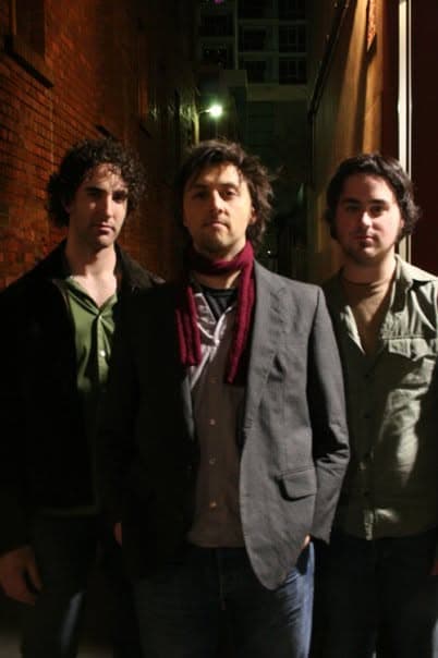 Three band members pose in a dim brick alleyway lit by a green light, the central one in a scarf and grey blazer.