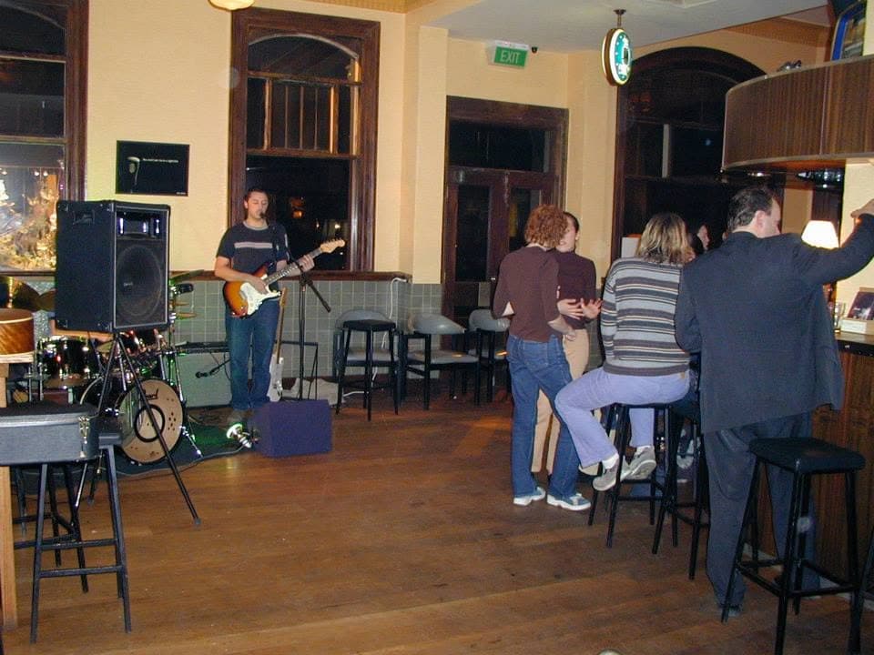 A band member plays electric guitar and sings into a mic in a wood-panelled pub while a few people sit and stand at the bar nearby.