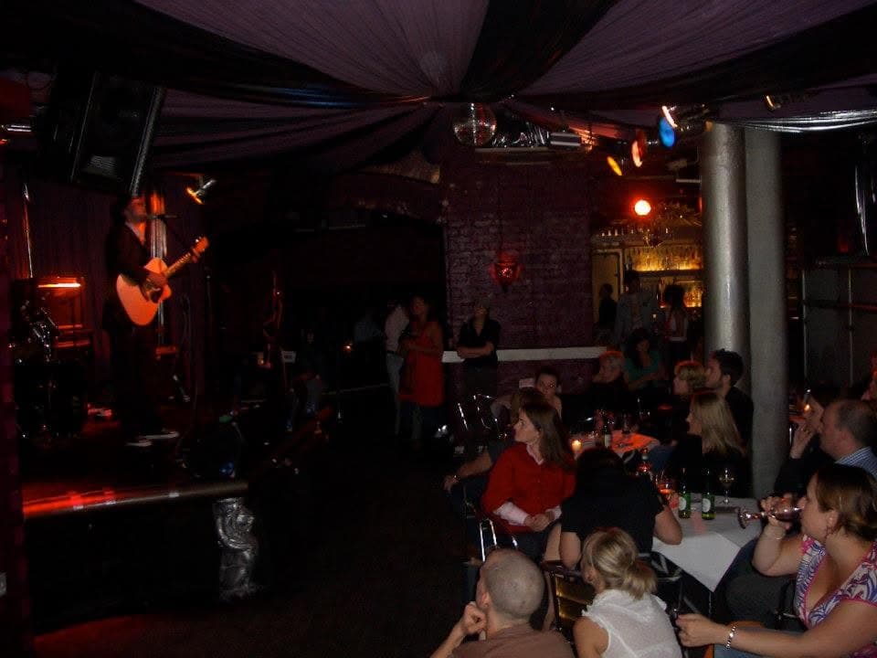A bearded musician plays acoustic guitar and sings on a club stage while a crowd watches from candlelit tables and the bar.