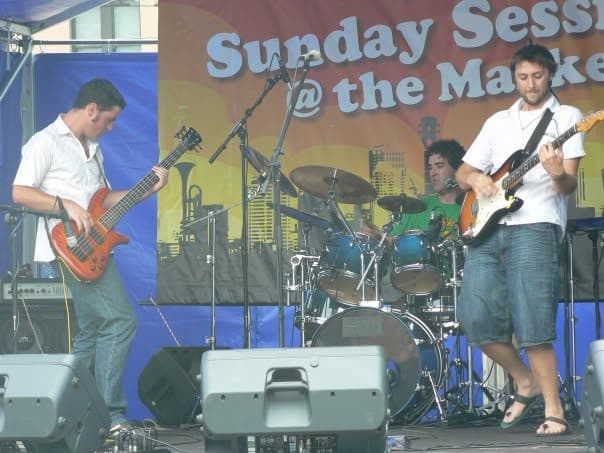 The band plays an outdoor daytime festival set under a 'Sunday Sessions @ the Market' banner, with Liam on bass at left, the drummer in green behind the kit, and a guitarist at right.