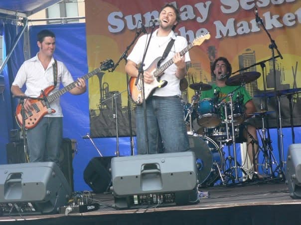 The trio plays a sunny outdoor gig with Liam on bass, Ben singing and playing guitar center, and the drummer mid-shout behind a blue kit.